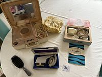 Photo showing four vintage personal care items: beige Remington device with mirror and speaker, cream-colored corded massager in open box with manual, gold foil pouch, and black paddle brush.