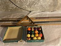 Four wooden pool cues laid out with an open box of vintage billiard balls and wooden triangle rack
