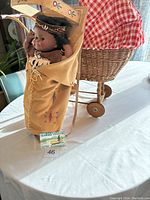 Image showing the Ojibaw crafts doll in traditional tan leather outfit with beadwork and feather decorations, positioned next to the wicker stroller on a white surface.