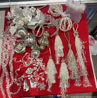 Overview photo showing arrangement of silver and white glass and acrylic Christmas ornaments including balls, tassels, flowers, and bead garlands on red background.