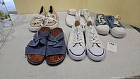 Lot of six pairs of ladies shoes and sandals on table including blue velcro sandals, white slip-on sandals, blue sneakers, cream shoes with lace front, beige canvas sneakers and beige slip-on shoes.