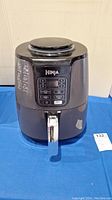 Front view of black Ninja air fryer on blue cloth with silver handle and control panel visible.