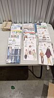 Wide view of numerous sewing patterns arranged on a table, including multiple brands and styles for ladies, children, and upholstery.