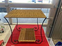 Front view of the wicker and metal side table showing wicker top and lower shelf with black metal scroll legs.