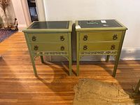 Front view of two matching antique side tables with green painted wood and dark green leather inlay tops with gold borders. Both have two drawers with decorative brass handles and carved aprons.