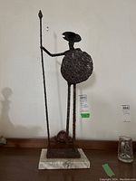 Front view of rough textured metal Don Quixote statue holding spear and shield on stone base, showing height and rust.