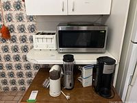 Photo showing LG microwave, white 4-slice toaster, coffee grinder, personal blender, hand mixer, and black coffee maker on kitchen counters and table.