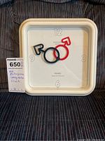 Full front view of the clock laid on fabric surface showing square white frame and unique overlapping male (black) and female (red) gender symbol hands on white face with minimal numeric markers.