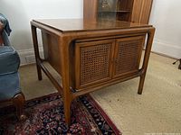 Side angle view of wooden mid-century modern side table showing rounded legs, rattan paneled double doors, and water stain on top.
