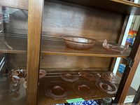 Wide view of wooden display cabinet shelves showing multiple pieces of pink depression glass including plates, candy dishes, and serving bowls arranged on two shelves.