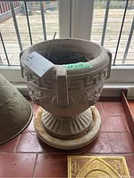 Front angled view of heavy concrete planter with grapevine motif and spinning base on tiled floor.