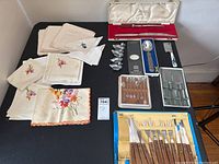 Overview of entire lot displayed on black table including napkins, teaspoons, cheese knives, Wostenholm carving set in case, and wood-handled utensils.
