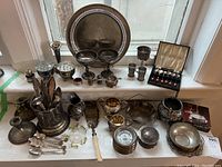 Wide view of lot items including trays, flower frogs, cutlery, goblets, coasters, pitcher, and other silver plated pieces on windowsill