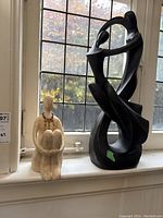 View of both statues together on a windowsill. Beige seated figure with bead necklace and taller dark wooden dancing couple statue with base crack.