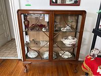 Front view of vintage wooden display cabinet showing two glass shelves with various items inside, wooden frame with decorative lattice on glass doors, cabriole legs visible.