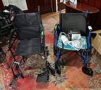 Two collapsible wheelchairs: left black fabric with metal frame, right blue Drive Blue Streak with lumbar pad and floral cushion. Accessories on blue chair seat include sleep machine controller and speakers.