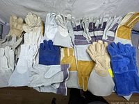 Various pairs of welding gloves in beige, blue, white, and gray colors with leather and fabric parts spread out on a flat surface.