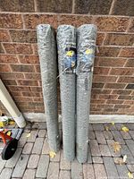 Three rolls of chicken fencing standing vertically against a brick wall, fully wrapped in plastic packaging.