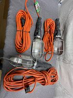 Three orange extension cords with built-in trouble lights placed on a gray surface. The lights have metal cages and hooks.