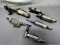 Photo showing 6 pneumatic air tools on grey fabric surface, various sizes and styles, used condition