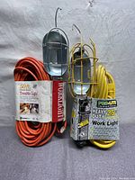 Photo showing pair of trouble light extension cords - one orange 50 ft and one yellow 25 ft, each with metal cage hand work light