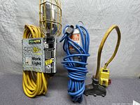 View showing the three items included in the lot: yellow Pro-Lite heavy duty 25 ft work light extension with cage, blue trouble light extension cord, and yellow GFI plug extension cord.