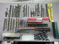 Photo showing a wide assortment of steel drill bits of varied sizes and types laid out on a surface, including some in plastic tubes and a Ryobi packaged bit.