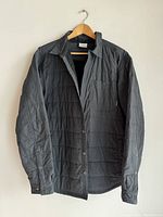 Full front view of black insulated men's overshirt on wooden hanger showing button closure and chest pocket.