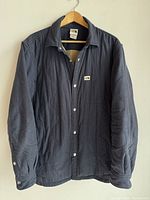 Front view of navy insulated overshirt hanging on wood hanger showing snap button front and collar.