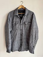Full front view of the grey Roots insulated overshirt hanging on wooden hanger showing buttons and chest pockets.