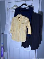 Four womens tops hanging on hangers on a door, including a charcoal gray blazer, white knit sweater, navy blue jersey top, and yellow plaid shirt