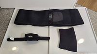 Set of three black medical support devices displayed on a white surface: large belt brace, wrist brace, and sleeve brace.
