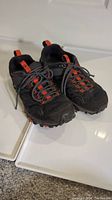 Pair of men's Merrell hiking shoes, black with red accents, size 8.5, shown from the front and top angles on a white surface with a carpet floor background.