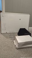 Photo of whiteboard leaning against wall with black metal file organizer, clear plastic file organizer, and three packs of white paper in front of it on carpeted floor.