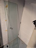 Thick, heavy glass sheet leaning against a door and wall in a corner, clear and rectangular with bevelled edges visible