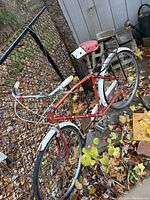 Full side view of vintage red glider bike leaning against chain link fence outside on leaves covered ground.