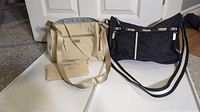Photo showing cream crossbody bag with attached wallet and black LeSportsac crossbody bag on a white surface in front of door.