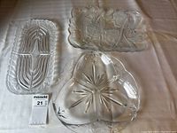 Three clear glass serving trays displayed on a white cloth. One tray is rectangular with a segmented fan-like pattern, another rectangular tray has raised floral designs, and the third is heart-shaped with a starburst design and scalloped edges.
