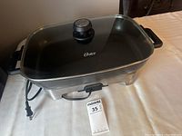 Front and side view of large Oster electric heating tray on a table, showing stainless steel sides, black handles, power cord, and glass lid with vent knob.