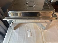 Large rectangular stainless steel buffet warming tray with lid and handle, elevated on four legs.