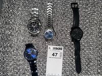 Top-down photo showing four men's watches placed on a woven textured surface: two with silver metal bands, one black leather band watch, one black metal band watch. Watch faces are a mix of black and blue dials.