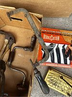 Box containing various antique hand tools such as hand drills and saws with wooden handles, a brass fireplace tool holder, and a boxed set of new BeanzCutter cutting tools.