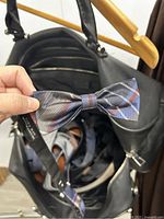 Photo of a plaid patterned men's bow tie held above the interior of the black leather duffle bag showing ties and belts inside.