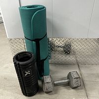 Photo showing the teal yoga mat rolled and strapped, TriggerPoint black foam roller, and a 30lb hexagonal dumbbell placed on the floor.