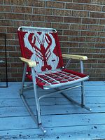 Side angle showing red and white woven lobster motif, metal frame, and beige armrests seated on a blue deck with a brick wall background.