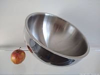 Large commercial stainless steel serving bowl shown angled to highlight reflective surface and size, apple for scale.