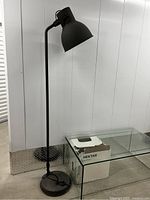 Full view of the Ikea dark grey floor lamp standing next to a clear glass table and box labeled Hektar.