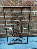 Front view of iron grate leaning against a brick wall on a blue wooden surface, showing full rectangular wrought iron design with curved elements.