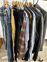 Men's clothing on rack showing denim jeans, pants, long sleeve shirts in plaid and solid, hoodies, and puffer vest.