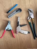 Photo showing a selection of hand tools on wooden floor including utility knives, crimping pliers, staple gun and adjustable pliers.